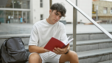 Cool young hispanic university student engrossed in reading a captivating book on the sunny city campus stairs, an embodiment of the relaxed yet serious college lifestyle. - obrazy, fototapety, plakaty