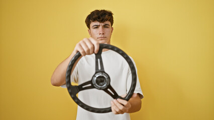 Handsome young hispanic teenager with a serious expression mastering the steering wheel as a relaxed driver, against an isolated yellow background.
