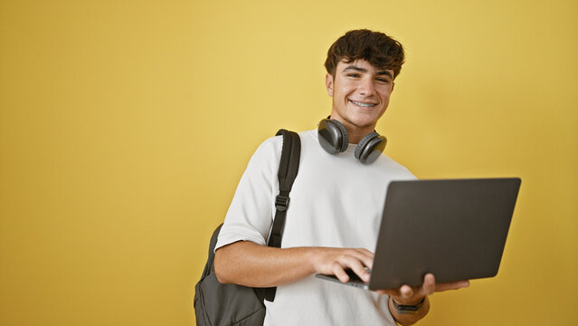 Cool young hispanic teenager enjoying life, smiling confidently with laptop & headphones. casual student guy sporting a backpack, isolated against a vibrant yellow wall.