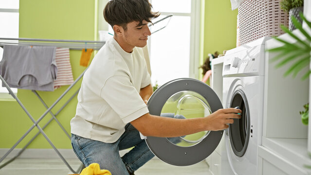 Smiling young hispanic teenager enjoying doing laundry, confidently inserting detergent bag into washing machine in laundry room