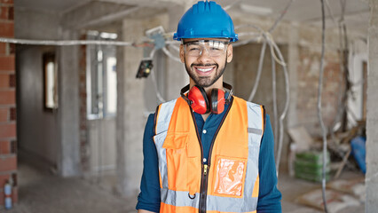 Young hispanic man builder smiling confident wearing hardhat at construction site