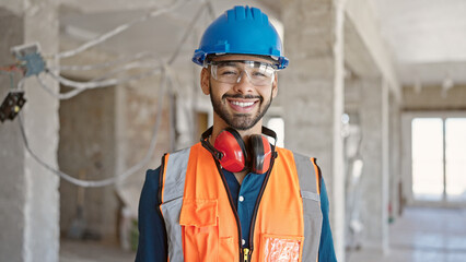 Young hispanic man builder smiling confident wearing hardhat at construction site