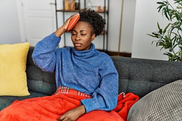 African american woman holding hot water bag on head sitting on sofa at home