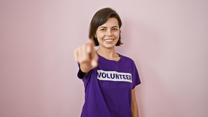 Smiling young volunteer, beautiful hispanic woman playfully pointing at you over isolated pink...