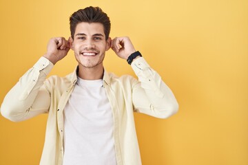Young hispanic man standing over yellow background smiling pulling ears with fingers, funny...