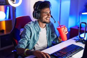 Young hispanic man streamer playing video game drinking beverage at gaming room