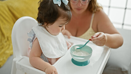 Mother and daughter together, loving family lifestyle, casually sitting at home during baby's lunch time, holding spoon and bib, concentrating on eating in the living room interior.