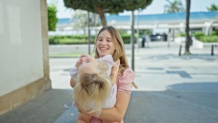 Fototapeta na wymiar Cheerful caucasian mother and daughter embracing a warm, loving hug, sitting on a city bench, laughing, and smiling together, filled with joy and happiness, outdoors on a sunny street.