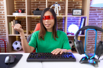 Middle age chinese woman wearing virtual reality glasses smiling with happy face looking and pointing to the side with thumb up.