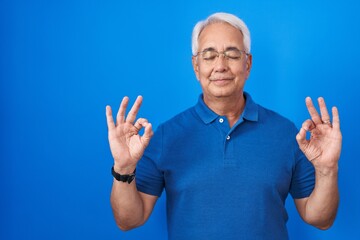 Middle age man with grey hair standing over blue background relax and smiling with eyes closed doing meditation gesture with fingers. yoga concept.