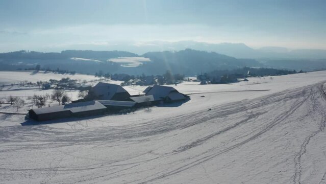 Perfect world in winter: A kid sledding down a hill in front of a farm, with the swiss alps in the background. Aerial drone footage in central europea