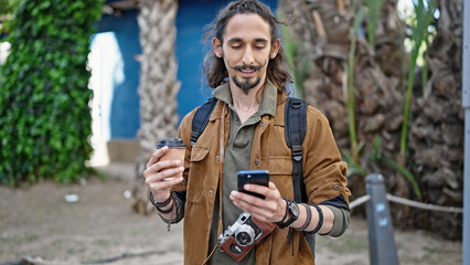 Young hispanic man tourist using smartphone holding coffee at park