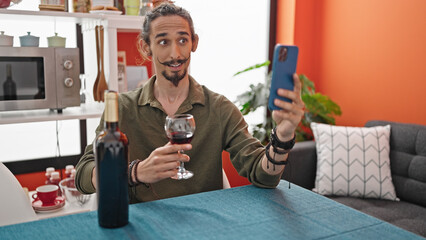 Young hispanic man drinking glass of wine having video call at dinning room