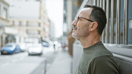 Serious, middle age man with grey hair, glasses, and a beard stands outdoors. as he looks up, his...
