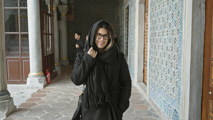 A smiling young woman with glasses, draped in a black shawl, stands by ornate tiles in istanbul.