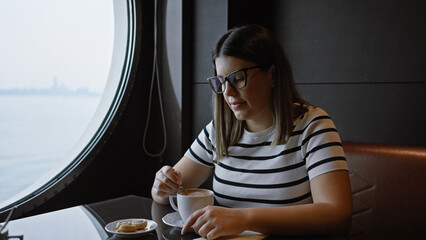 A young woman enjoys her coffee at a table by the window inside a cruise ship cafe.