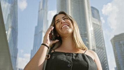 Fototapeta premium A smiling young woman talks on her phone in front of the towering skyscrapers of dubai under a blue sky.
