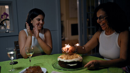 Two women, likely mother and daughter, celebrate indoors with a sparkler-topped cake and wine glasses on the table.
