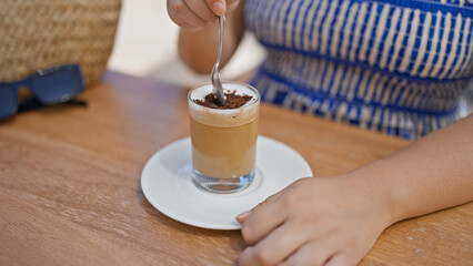 Young hispanic woman stirring coffee sitting on the table at sunny restaurant terrace