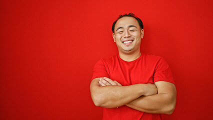 Laughing handsome young asian man exudes confidence, standing with arms crossed over red background, embodying joy and positivity