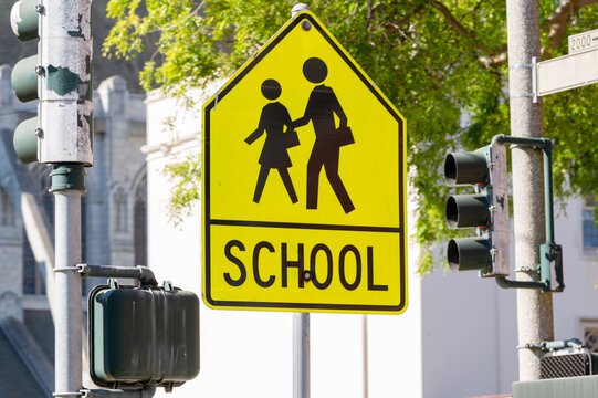 Yellow warning road sign for a school zone at a road intersection with a street traffic light in the background. Yellow School Zone Street Sign, Close-up. School Road Sign with Children, Foreground