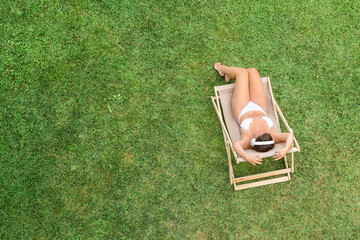 Woman in a white bikini in headphones with basket for picnic sitting on deck chair on the green...