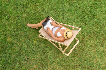 Woman in a white bikini sitting on deck chair with laptop on the green grass sunbathes at summer day. Top view, drone, aerial view. - 721527738