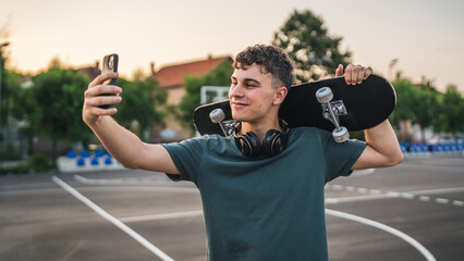 Fototapeta premium One man young adult caucasian teenager stand outdoor with skateboard on his shoulder and headphones posing at basketball court portrait happy confident wear shirt casual real person copy space