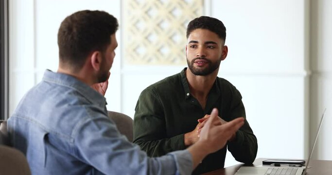 Young Moroccan businessman talk to male colleague, discuss new project sitting at desk in office, share ideas or business plan. Executive manager gives instructions new employee. Negotiations, meeting