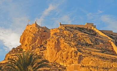 Naklejka premium Rocky mountain with Santa Barbara castle in the evening sun with blue sky and white clouds, in Alicante, Spain, Europe