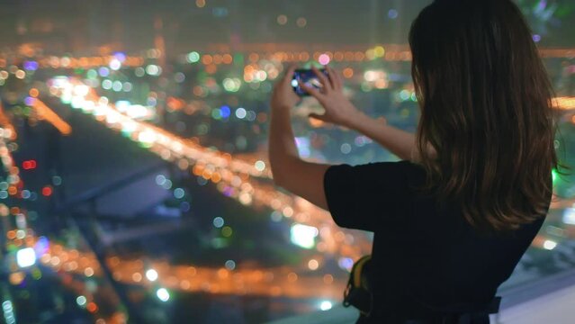 Girl enjoy night cityscape traffic road highway from top floor window. Woman make aerial city panorama shot on her mobile phone. Colorful center downtown, night lights bokeh. Close up, slow motion