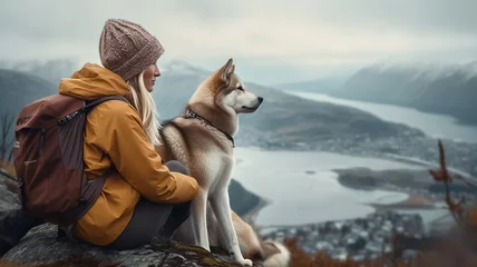  Cinematic image of a hiker girl sitting with husky at the top of the mountain with rocks, autumn trees and lake. Long shot of a beautiful scene in autumn from the top. Moody colors. © Loucine