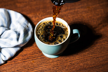 Black coffee being poured from a carafe into a small green teacup on a wooden table with a diner style towel nearby