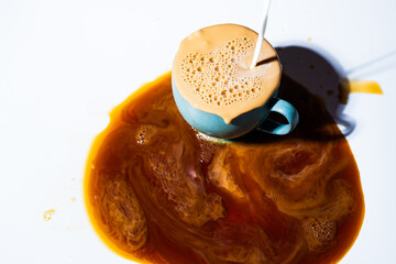 A green teacup filled to the rim with creamed coffee spilling out into a round puddle on a white table as more cream is poured in from above