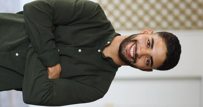 Vertical shot of happy middle-eastern young man in casual shirt standing indoors, posing with arms-crossed on chest. Profile portrait of promoted successful employee, high school or college student