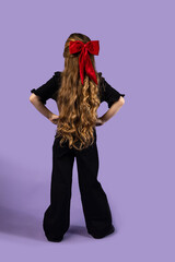 Full back shot of a little girl with long, wavy blonde hair wearing black clothes, hands on hips, isolated on purple background.