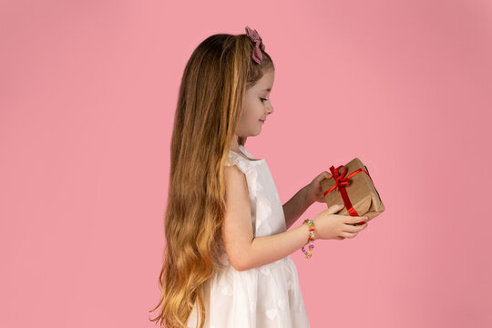 The picture of the little girl who is dressed in a white dress and holds a gift in her hand and is very excited about what is in it. picture on a pink background. The girl is beautiful with two