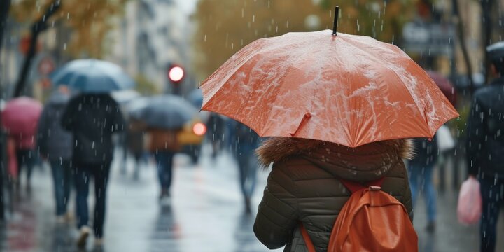 A woman is seen walking down the street with an orange umbrella. This image can be used to depict a rainy day or protection from the sun