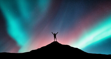 Northern lights and silhouette of a sporty man with raised up arms on the mountain peak in Norway....
