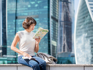 Smiling woman reads paper map. She is sitting on background of buildings with glass walls. Travel around city. Urban tourism. Modern architectural landmarks. - 721637523