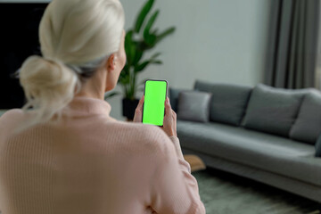 Over the shoulder shot of a person holding an phone with a completely chroma green screen. Mature woman in the living room