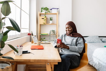 Happy young black woman in muslim headscarf using smartphone at home. Social media, youth lifestyle and technology concept. - obrazy, fototapety, plakaty