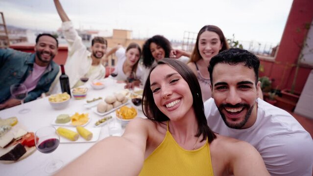 Excited young pretty woman recording selfie video for social media on rooftop. Cheerful female looking funny making faces with diverse friends to camera outdoors. People gathered celebrating lunch 