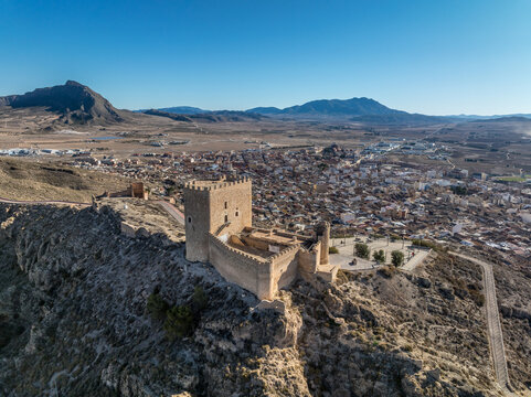 Aerial view of Jumilla medieval castle in Murcia Spain, on a hilltop, imposing irregular shape keep with four floors, crenelated battlements cloudy blue sky background