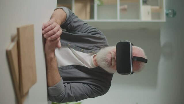 Vertical View.A Modern Old Man With Gray Beard in VR Glasses Examines Everything Around. An Elderly Man in VR Glasses Sitting at a Table at Home.VR to Help the Elderly.