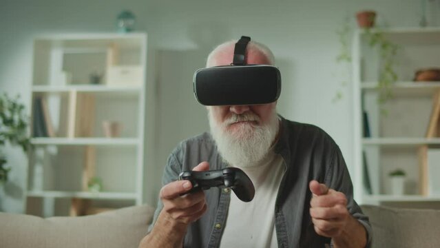 An Old Man in VR Glasses Plays Computer Games With a Joystick, Sitting in the Sofa and Eating a Popcorn.A Modern Elderly Man Plays PlayStation. Augmented Reality and Gaming for Seniors.