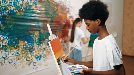 Closeup of african boy hand paint canvas with warm tone color while caucasian girl in casual outfit...