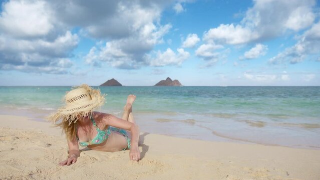 Love and happiness concept. Female traveler sunbathing Lanikai beach Mokulua islands view. Smiling tourist in fringed straw hat drawing heart in golden sand on Oahu beach. Young woman tanning on beach