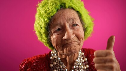 Slow motion funny view of mature elderly woman, 80s, having giving OKAY gesture, wearing green wig or hat isolated on pink background.