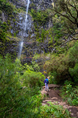 Obraz premium Female tourist with backpack hikes torwards a high waterfall at a rock wall. Lagoa do Vento waterfall, Madeira Island, Portugal, Europe.